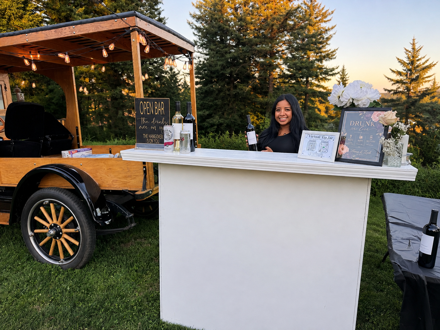Zaira behind her white mobile bar at an outdoor event, with trees and string lights